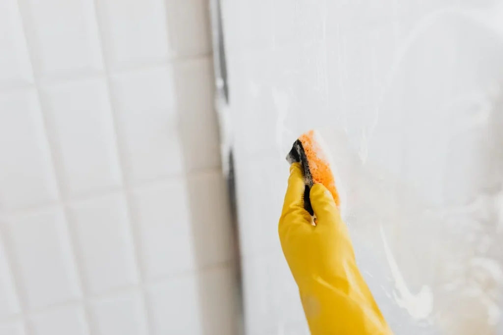 main gantée lavant la cabine de douche en verre avec une éponge
