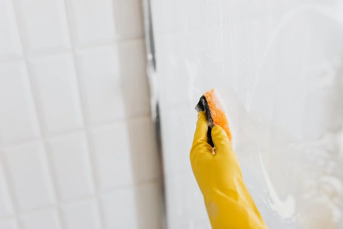 main gantée lavant la cabine de douche en verre avec une éponge